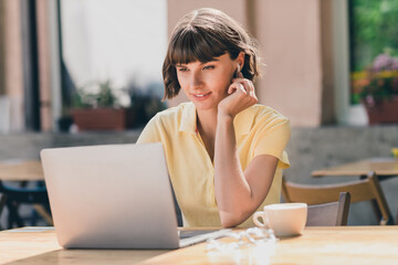 Portrait of attractive focused cheery girl using laptop sitting at cafe working project start-up on fresh air outdoors