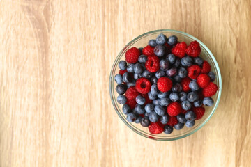 Glass bowl filled with raspberries and blueberries on wooden table. Top view.