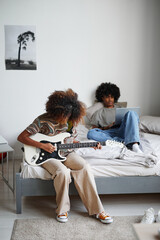 Vertical full length portrait of two teenagers at home, focus on young African-American woman playing electric guitar