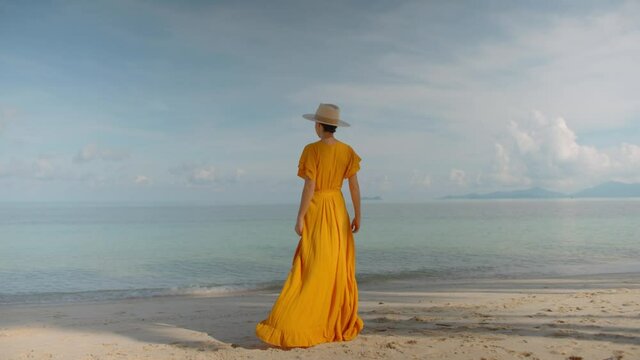 Back shot of nice woman in yellow long dress walking to edge of sea water enjoying windless scenery