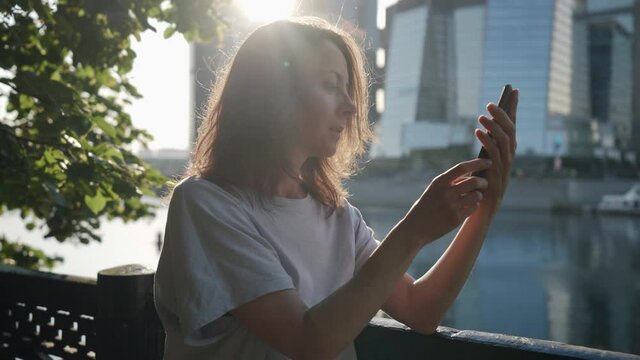 a woman leans on the fence and attentively looks at the phone screen on the embankment against the background of modern city buildings. contour light. the camera is moving. side view