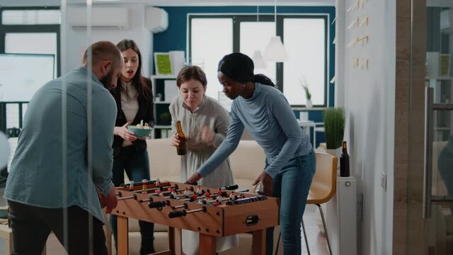 Woman winning game of soccer at foosball table while playing with man at office, having fun after work. Multi ethnic colleagues playing for entertainment, enjoying beer drinks.