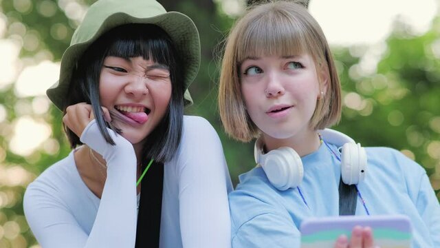 Two smiling multinational friends taking selfie with tongue sticking out on the phone outdoors