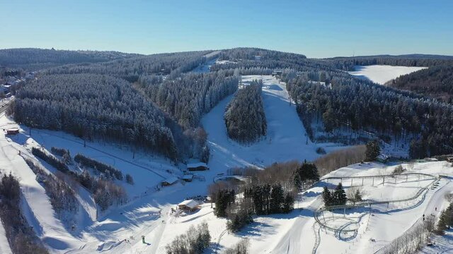 Aerial view of winter sports slopes at ski lift carousel Winterberg. Toboggan slope and ski slopes between snow covered spruce forests. Camera flies down.