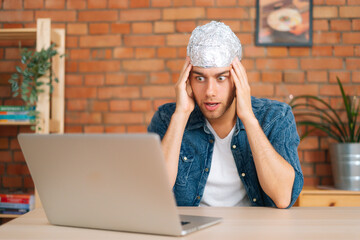 Portrait of scared afraid male conspiracy theorist wearing aluminum foil protect brain watching shocked online video content using laptop computer, sitting at desk in living room with modern interior