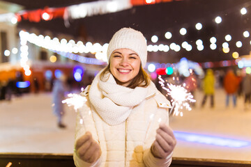 Smiling young woman wearing winter knitted clothes holding sparkler outdoors over snow background. Christmas holidays.