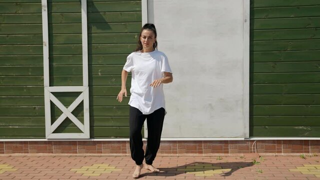 barefoot brunette in black pants and a big white T-shirt dances against a green wall on the street on a sunny day. she tramples, bends knees and makes a wave with legs. the general plan