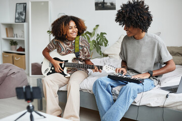 Portrait of two African-American young people playing music at home and recording video or...