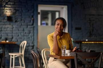 African american woman using a smartphone and drinking coffee while sitting in a cafe