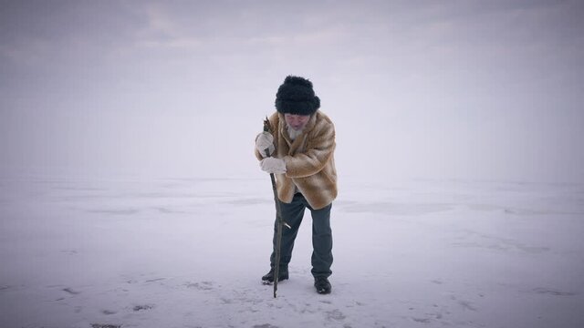 Wide shot bearded senior man hitting stick on ice in slow motion standing on frozen ocean. Confident indigenous old guy fishing on arctic landscape