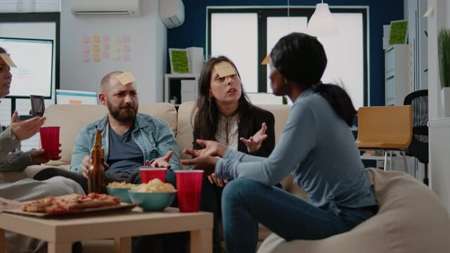 Multi ethnic group of workers playing charades to guess sticky notes after work for entertainment. Diverse colleagues enjoying guessing game play to do fun activity at celebration.