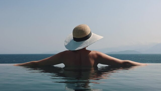woman with straw hat relaxing in infinity swimming pool with sea view at luxury resort. summer vacation