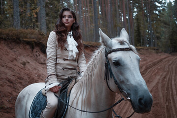 A young beautiful brunette rider in an elegant retro suit riding a white horse in a forest area
