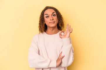 Fototapeta premium Young latin transsexual woman isolated on yellow background winks an eye and holds an okay gesture with hand.