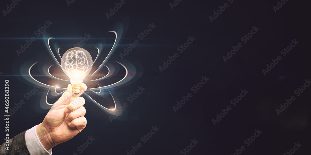 Leinwandbilder close up of hand holding abstract light bulb on dark wide background with digital brain and atom out