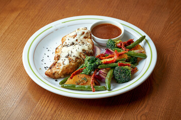 Baked chicken breast with cheese and boiled vegetables, broccoli in a white plate on a wooden table