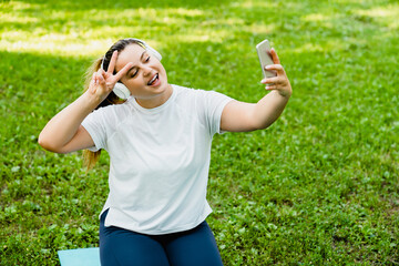 Fitness strong muscular fit plump plus-size caucasian woman athlete taking selfie photo image, having video call conversation with trainer in headphones, posting on social media outdoors in park