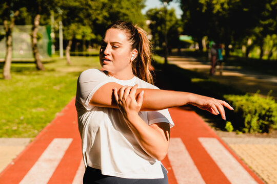 Fit young plump plus-size body positive woman athlete stretching her arm before yoga class training losing weight in fitness outfit in stadium