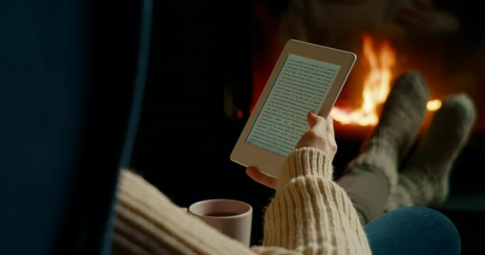 Woman in Sweater Reads eBook on electronic Reader relaxing at Fireplace in Cozy Room at Winter Night. Christmas Seasonal Leisure or Comfortable Warm Autumn Evening Rest. 4K slider pan medium shot