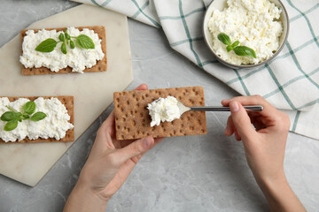 Woman spreading cottage cheese onto crispy cracker at grey marble table, top view