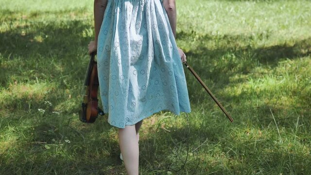 A girl in a dress walks with a violin in a city park.