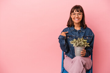 Young mixed race woman sitting on a chair holding a plant isolated on pink background person pointing by hand to a shirt copy space, proud and confident