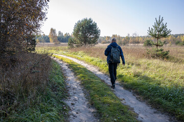 Obraz premium A tourist walks along a country road early in the morning. A man with a backpack on the background of a field illuminated by sunlight.