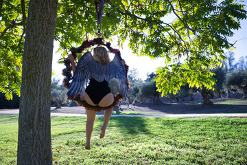 Young blonde woman in a black dress with angel wings sitting backwards on an aerial hoop hanging from a tree. Halloween concept, angels, fallen angel, aerial hoop, curvy girl. - obrazy, fototapety, plakaty
