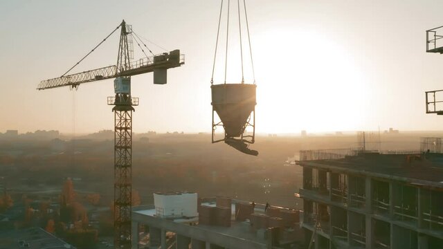 Aerial panoramic view of a concrete bucket hanging from a tower crane at a large construction site with high-rise residential buildings.