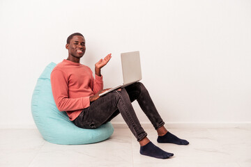 Young African American man sitting on a puff using laptop isolated on white background showing a copy space on a palm and holding another hand on waist.