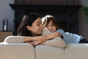 Excited 7s son boy and happy Asian younger mom playing and hugging on couch, messing up, laughing...