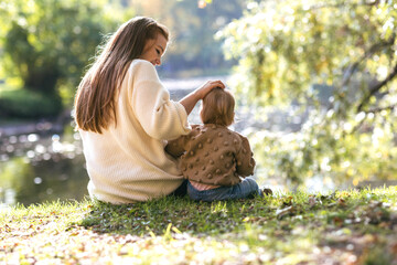 A young mother with a one-year-old baby in an autumn park hugs on the edge of the lake and smile. Filmed from the back