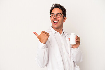 Young pharmacist mixed race man holding pills isolated on white background points with thumb finger...
