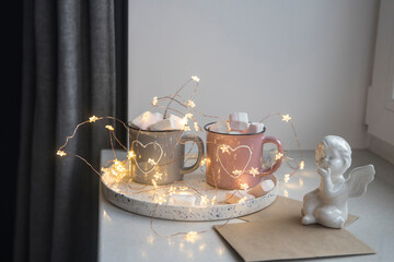 Apartment decorations. Gray and pink mugs with painted white hearts with cocoa, marshmallows on a tray on the window, composition decorates the interior on the eve of Christmas. Copy space