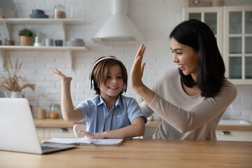 Excited gen z schoolkid in headphones giving high five to happy mom at laptop. Mother helping son...