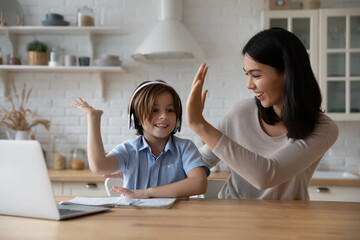Excited gen z schoolkid in headphones giving high five to happy mom at laptop. Mother helping son to do school home work, giving praise and support for studying achievement. Parenthood, homeschooling