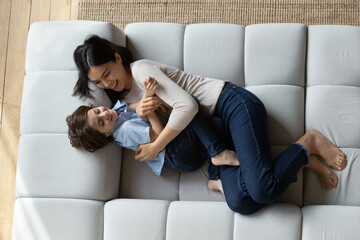 Happy young Asian mom tickling and cuddling laughing son boy on sofa. Mum and little 7s kid...