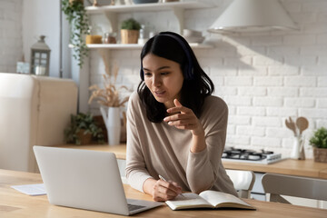Positive Asian student in headphones attending virtual class, online training conference, looking at laptop screen, talking on video call, watching learning webinar, writing notes, studying from home