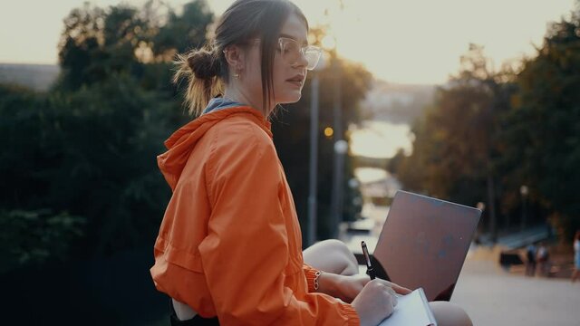 The young, thoughtful journalist looks for the right words for the article, when she finds it she continues to write. Dressed in a bright orange blouse, hair caught in a bun, positioned on the park
