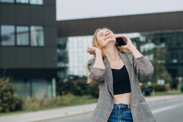 Portrait of young happy blond woman with phone walking on the street in the city. Technology or people concept. Excited girl laughing on bulding background.