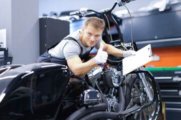 Fototapeta na wymiar Young car mechanic is diagnosing a motorcycle and holding clipboard and thumbs up
