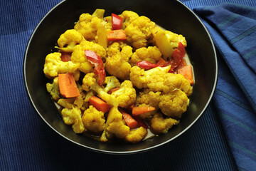Curried cauliflower with carrots and red sweet pepper in a black bowl on a blue background