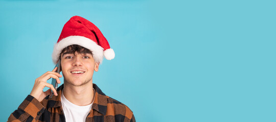 portrait of teenage boy talking on mobile phone wearing santa claus hat isolated on blue background