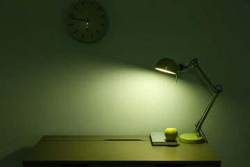 Glowing lamp, apple and book on table in dark green room