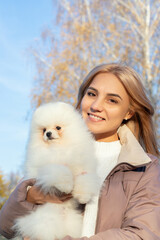 young beautiful woman holds white pomeranian dog with fluffy fur in her arms in city park