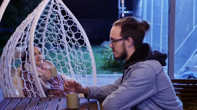 The concept of family dialogue: Mom is drinking tea in a cozy street chair and talking with an adult son in the gazebo of her own home. Guy with glasses with a beard ius