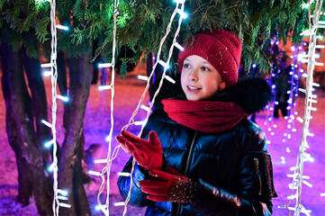 beautiful young girl in red cap and scarf stands against background of  Christmas tree and  lights...