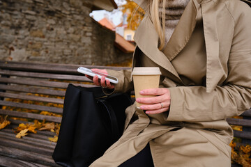 Girl with a smartphone and a cup of coffee sits on a bench in an autumn park. A woman is sending a text message or using an app on her mobile phone. Hot drink in a paper cup for a cozy walk
