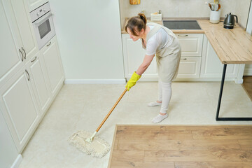 A woman in an apron cleans the floor with a mop in a home kitchen