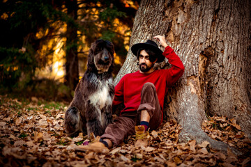 a young man, dark-haired, in a red sweater and hat,sitting under a tree with a pet dog,in the afternoon in the autumn park